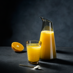 Orange juice in a glass and pitcher on a dark surface with an orange half.